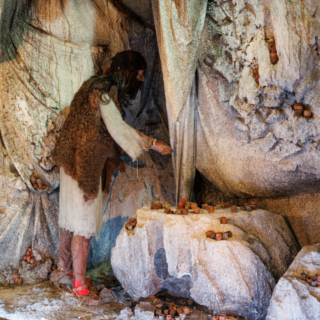 Una figura umana a grandezza naturale, rappresentante un individuo dell'età del Bronzo con lunghi capelli e vestiario in tessuto chiaro e pelliccia, si china all'interno di una grotta artificiale. La figura interagisce con piccoli manufatti cilindrici color terracotta, simili a vasetti, disposti su una roccia e presenti anche nelle nicchie delle pareti rocciose circostanti.