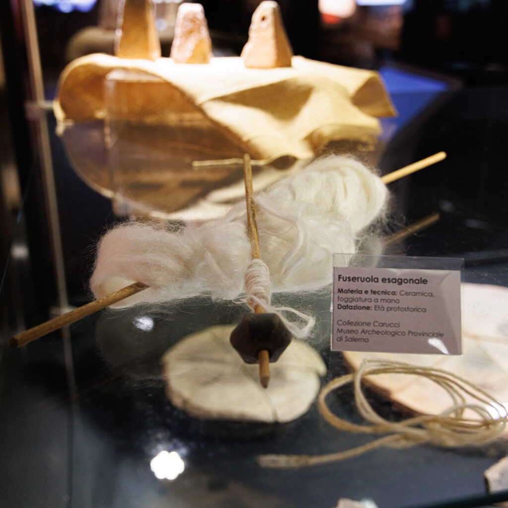 Reperti archeologici legati alla tessitura in una teca museale: in primo piano, un fuso in legno con fibre bianche avvolte e una fuseruola esagonale in ceramica scura alla base. Sullo sfondo, un tessuto con alcuni pesi da telaio conici. Un'etichetta indica: 'Fuseruola esagonale, Ceramica, Età protostorica, Collezione Carucci, Museo Archeologico Provinciale di Salerno'.